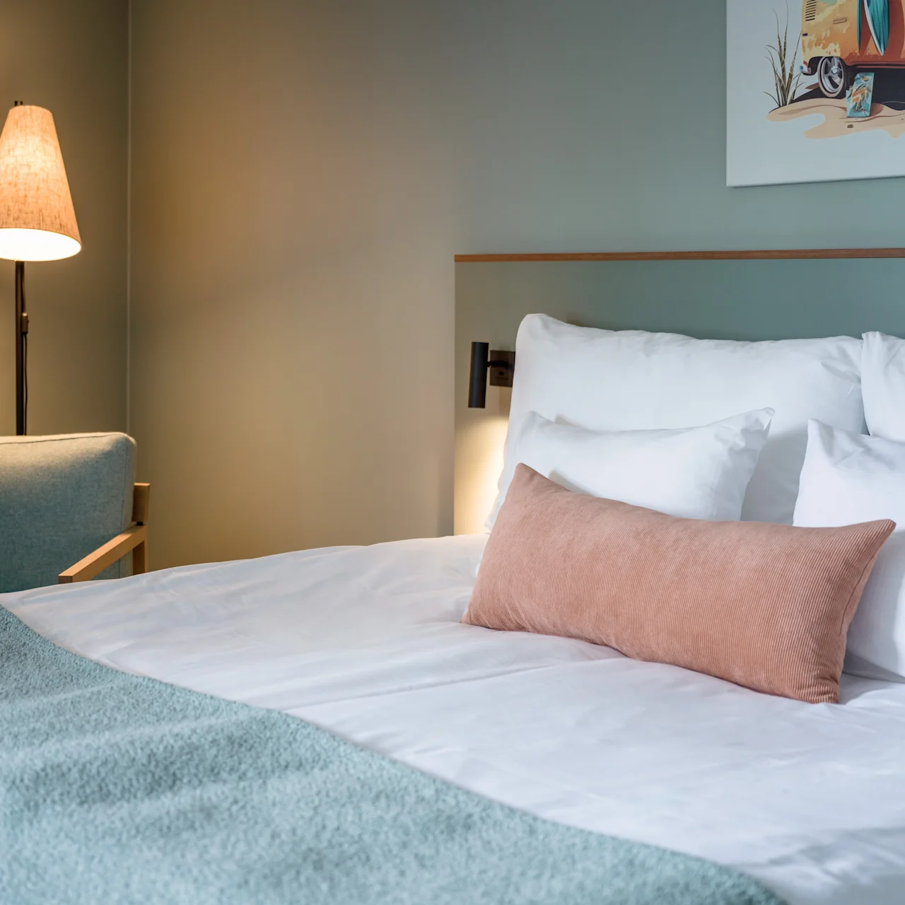 A neatly made bed with white and beige pillows stands in a serene room, accompanied by a floor lamp and modern armchair. A colorful painting decorates the muted walls.
