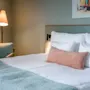 En pent oppredd seng med hvite og beige puter står i et rolig rom, akkompagnert av en gulvlampe og moderne lenestol. Et fargerikt maleri dekorerer de dempede veggene.