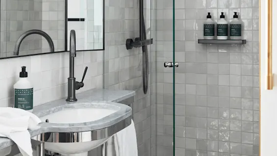 Sink with a black faucet stands in a tiled bathroom. Three soap bottles rest on a glass shelf in the shower. A round mirror and towel complete the setting. Text: "SKOG" and "HAND WASH".