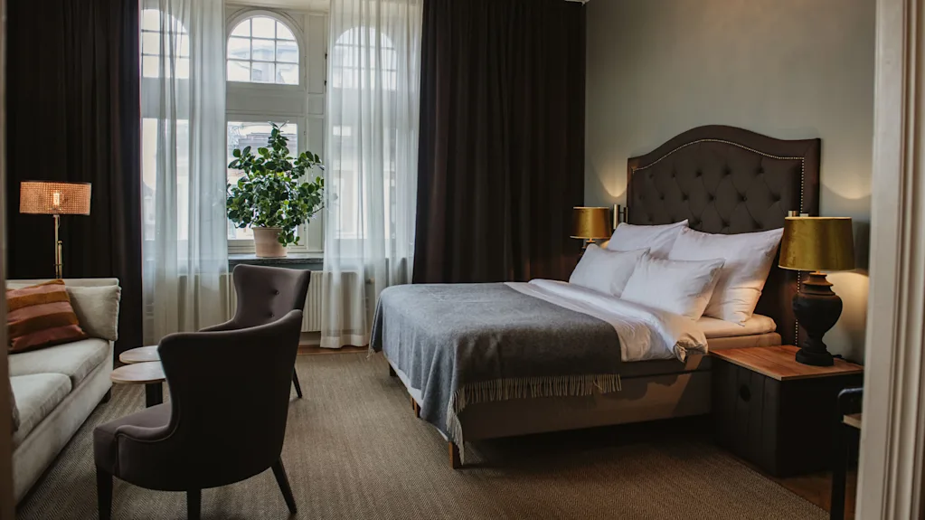 A bed with white linens and a gray blanket is flanked by lamps on wooden nightstands; an armchair and sofa sit nearby, with a large window and curtains in the background.  