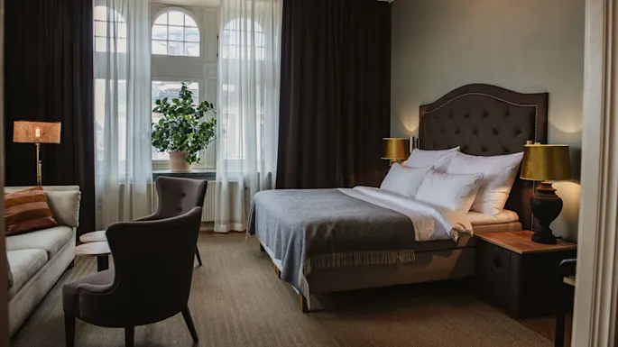 En seng med hvitt sengetøy og et grått teppe flankeres av lamper på trebenker; en lenestol og sofa står i nærheten, med et stort vindu og gardiner i bakgrunnen.