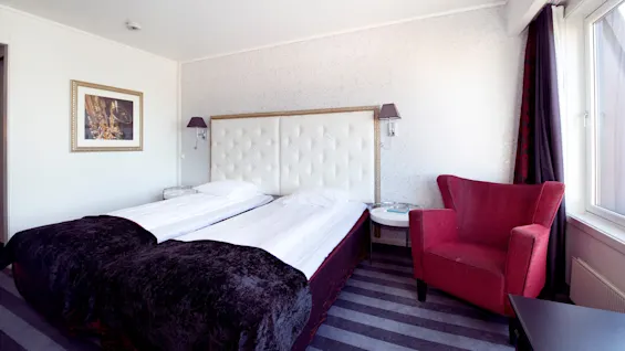 A neatly made twin bed anchors a hotel room with a framed picture on the wall, a red armchair, and striped carpet.