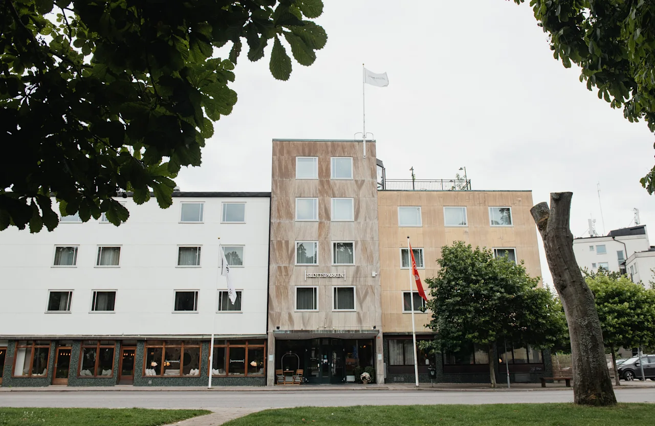 A large building with a sign reading SLOTTSSPARKEN, featuring different facades and flags, framed by green trees.