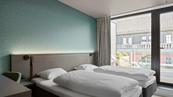 A neatly made twin bed sits in a modern room with teal wallpaper, beside a large window overlooking a building with tiled roofs.