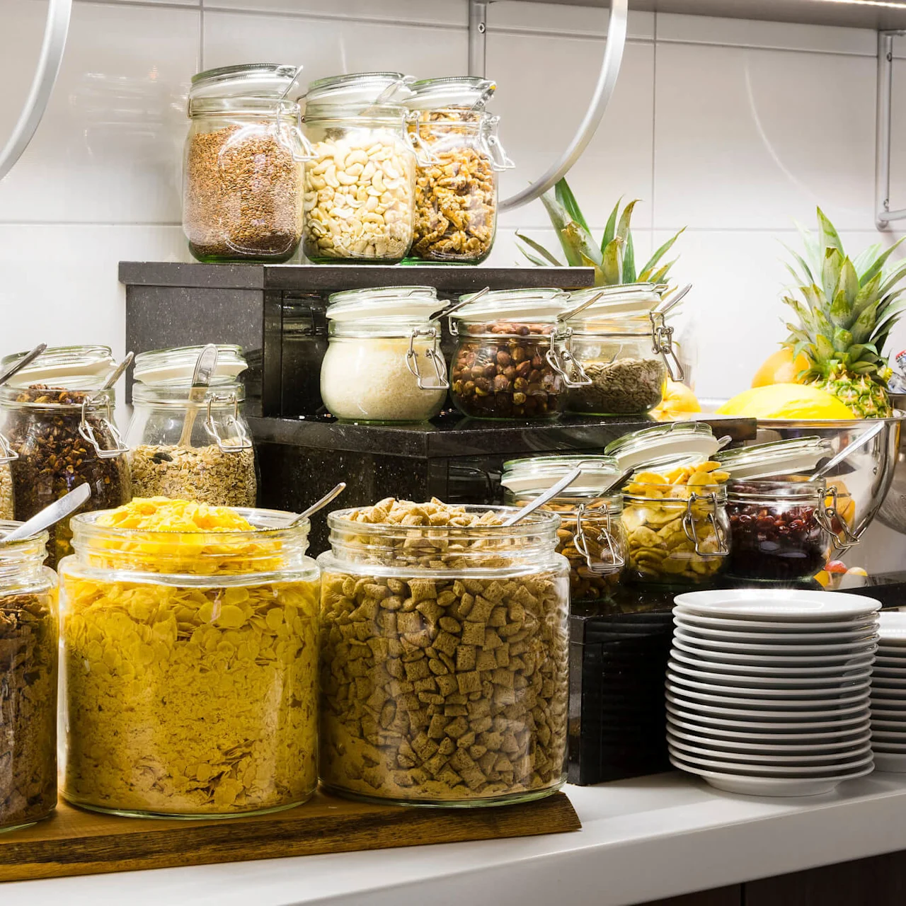 Ulike frokostblandinger i klare glasskrukker på en buffet med frukt og stablede tallerkener.
