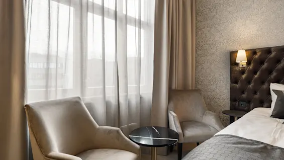 Et hyggeligt hotelværelse med to komfortable lænestole, et lille bord og en seng ved et vindue med lette gardiner.