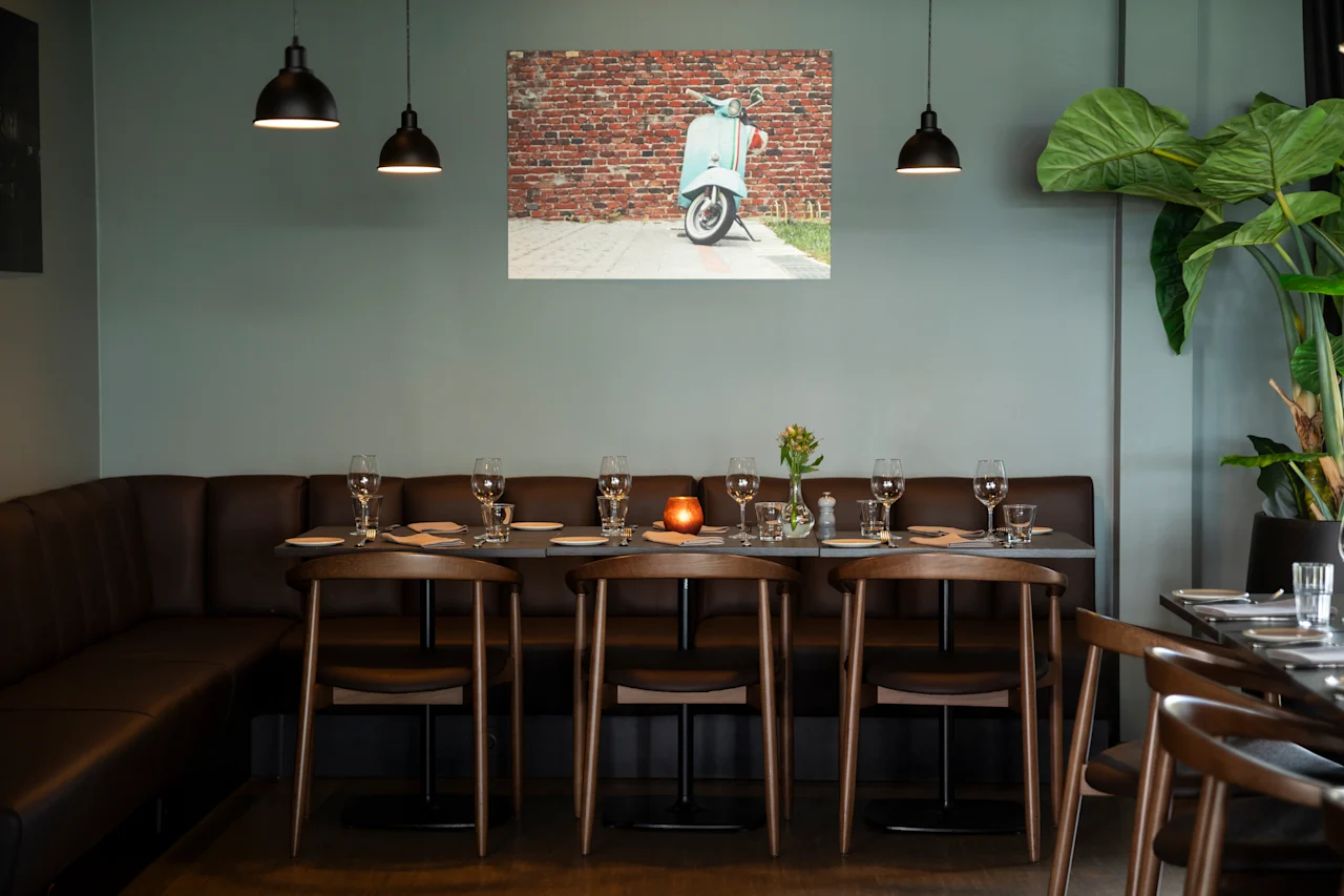 Et koselig restaurantinteriør med et langt spisebord dekket med glass og bestikk, en brun skinnbenk og trestoler. Et bilde av en scooter henger på veggen.