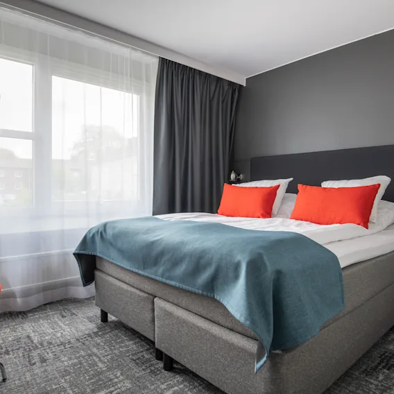 Ett modernt hotellrum med en bekväm säng, livfulla orange kuddar och en mysig läshörna vid fönstret.