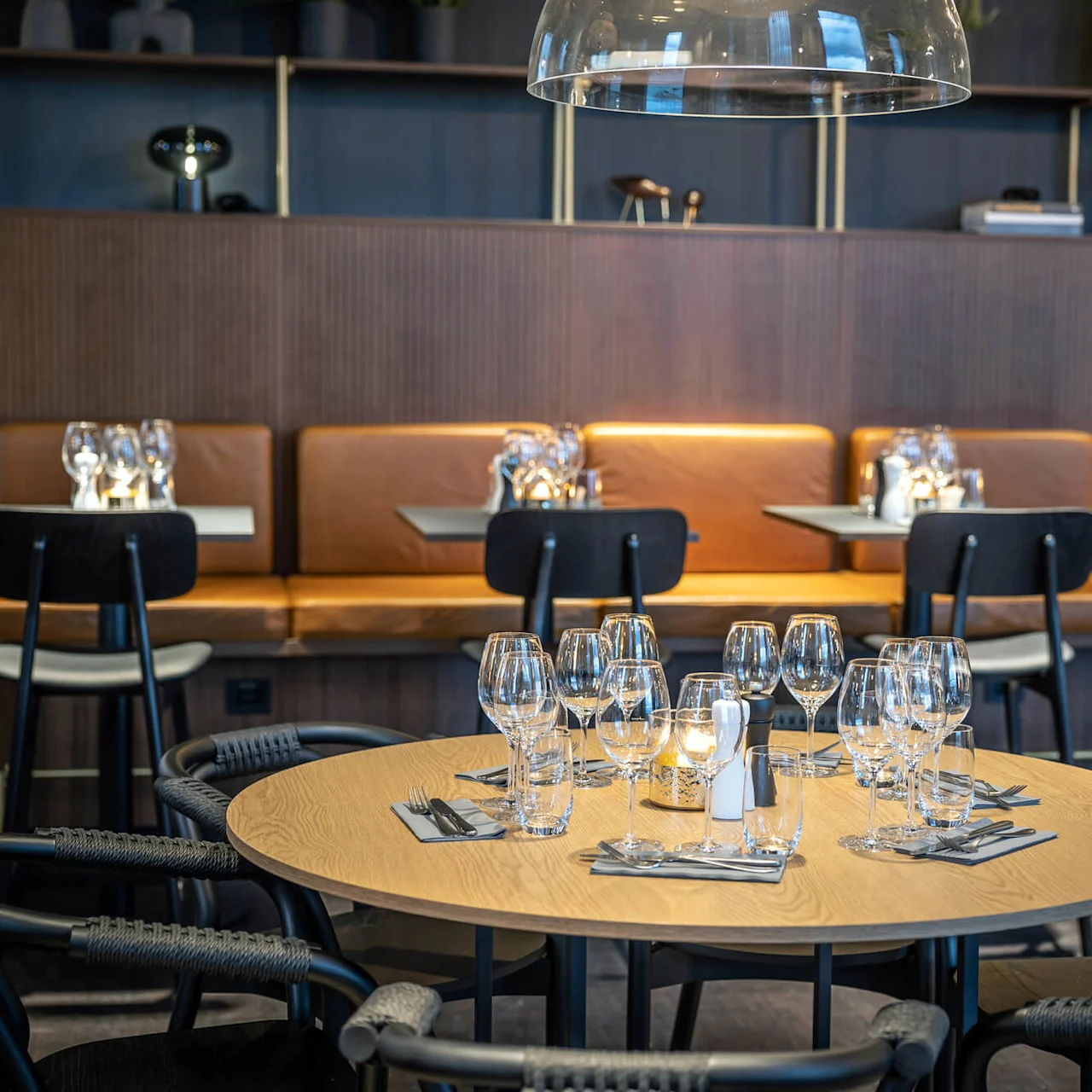 Et rundt bord dækket med mange vinglas og bestik, omgivet af moderne stole, placeret i et stilfuldt restaurantmiljø med brune læderstole mod væggen.