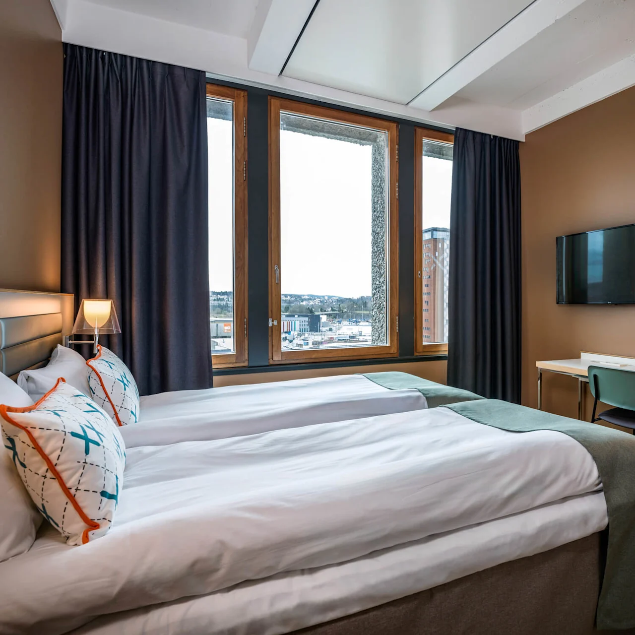 A neatly made bed with patterned pillows sits near a window with curtains, overlooking a cityscape. A desk with a chair and lamp faces a wall-mounted TV.