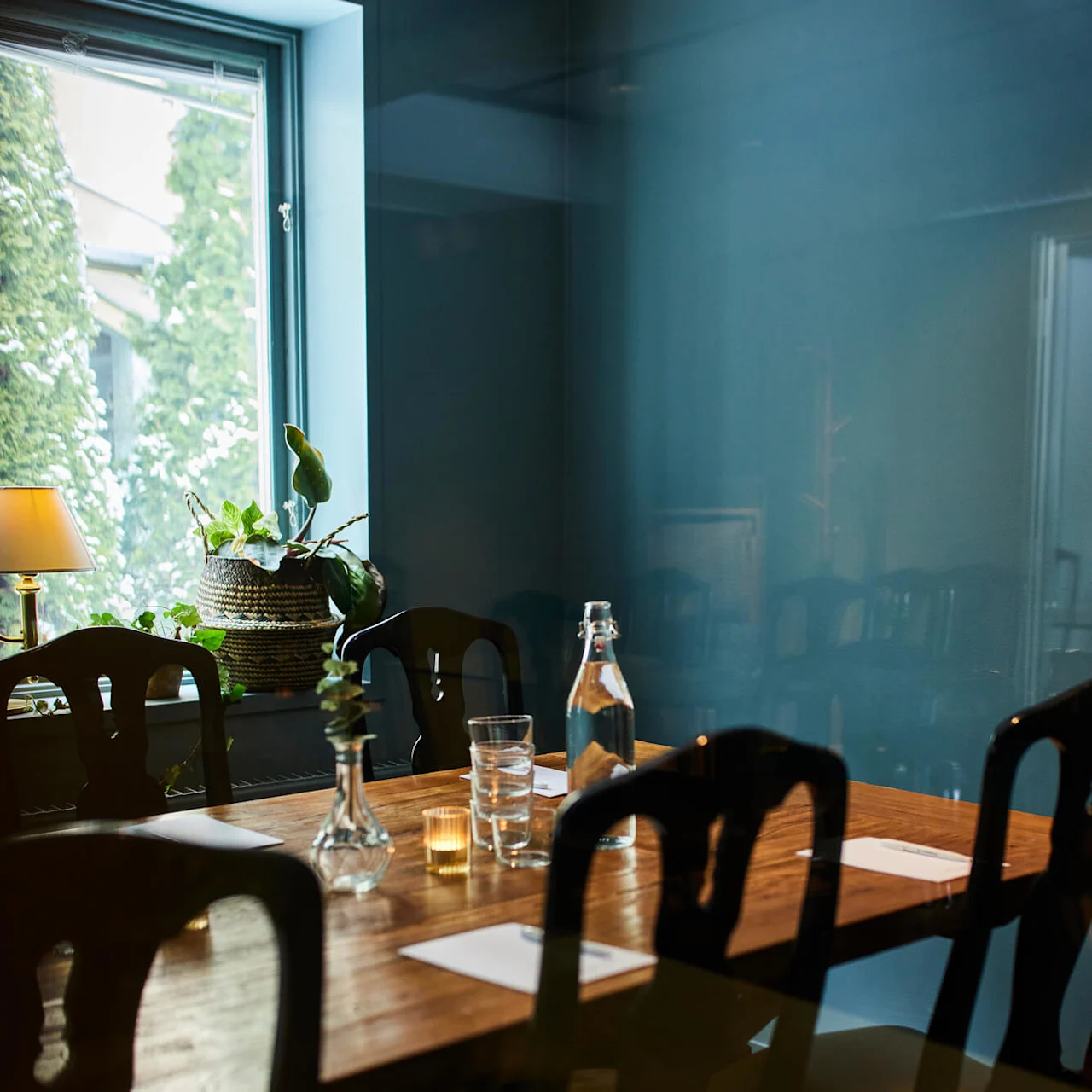 Et trebord dekket med notatblokker, glass og en vannflaske under myk belysning, omgitt av stoler i et rom med et vindu og potteplanter.