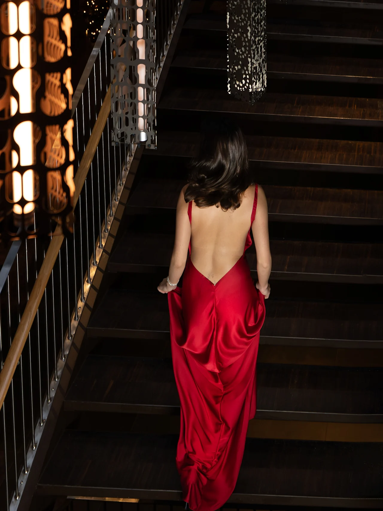 En kvinna i en rygglös röd klänning går uppför en svagt upplyst trappa, omgiven av intrikata metallräcken och dämpad belysning.