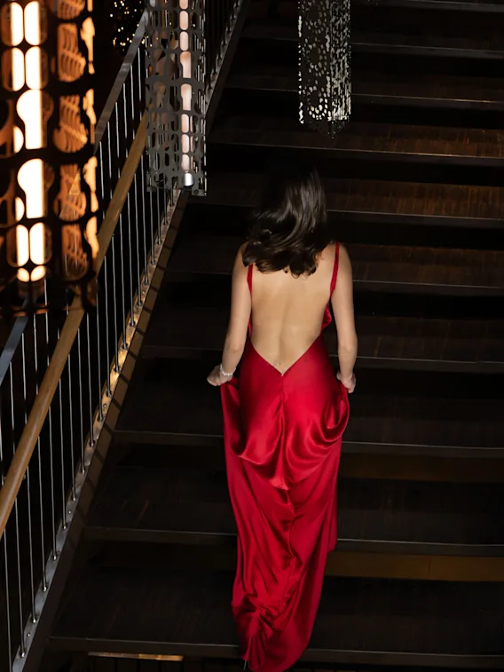 En kvinna i en rygglös röd klänning går uppför en svagt upplyst trappa, omgiven av intrikata metallräcken och dämpad belysning.