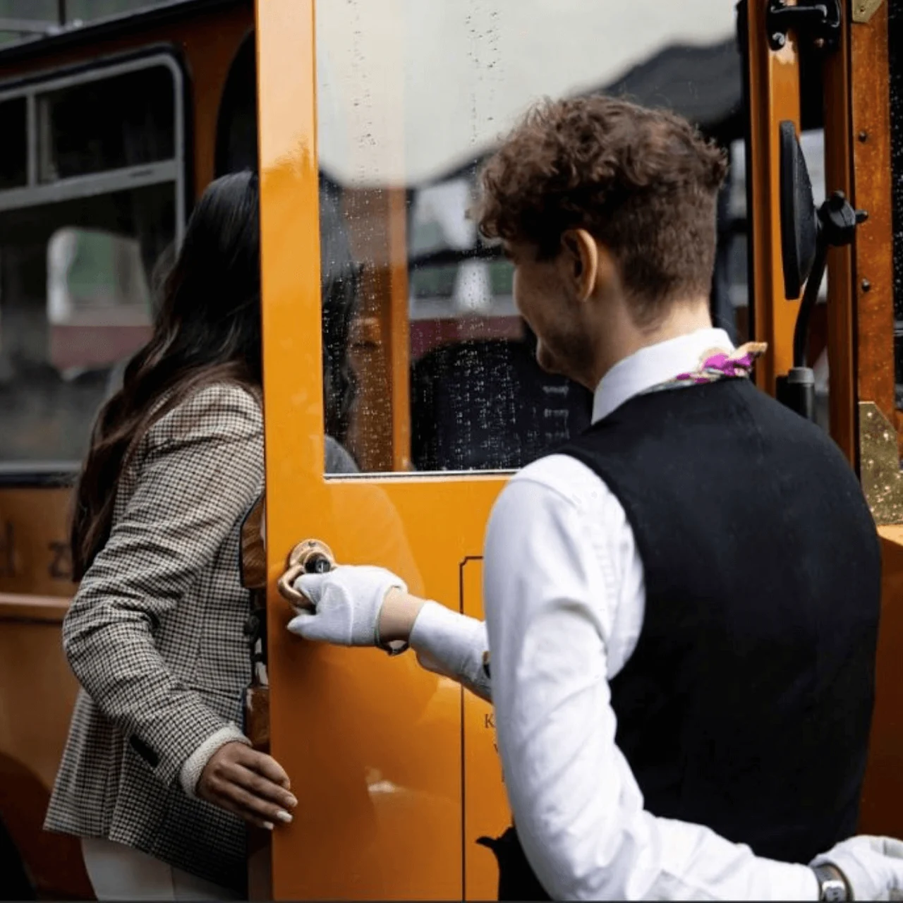 A woman enters an orange vintage bus assisted by a man in formal attire, outside a hotel. Another man stands nearby holding a hat. Text: "Kronenhof Kabelan Hotel".
