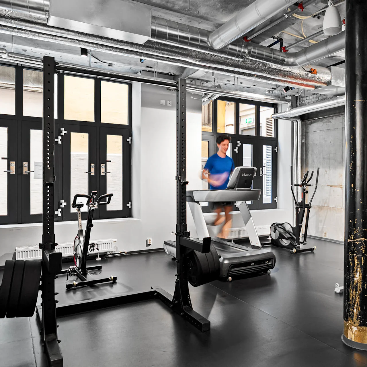 En person springer på ett löpband i ett modernt gym med svartvit inredning, omgiven av olika träningsredskap och stora fönster.