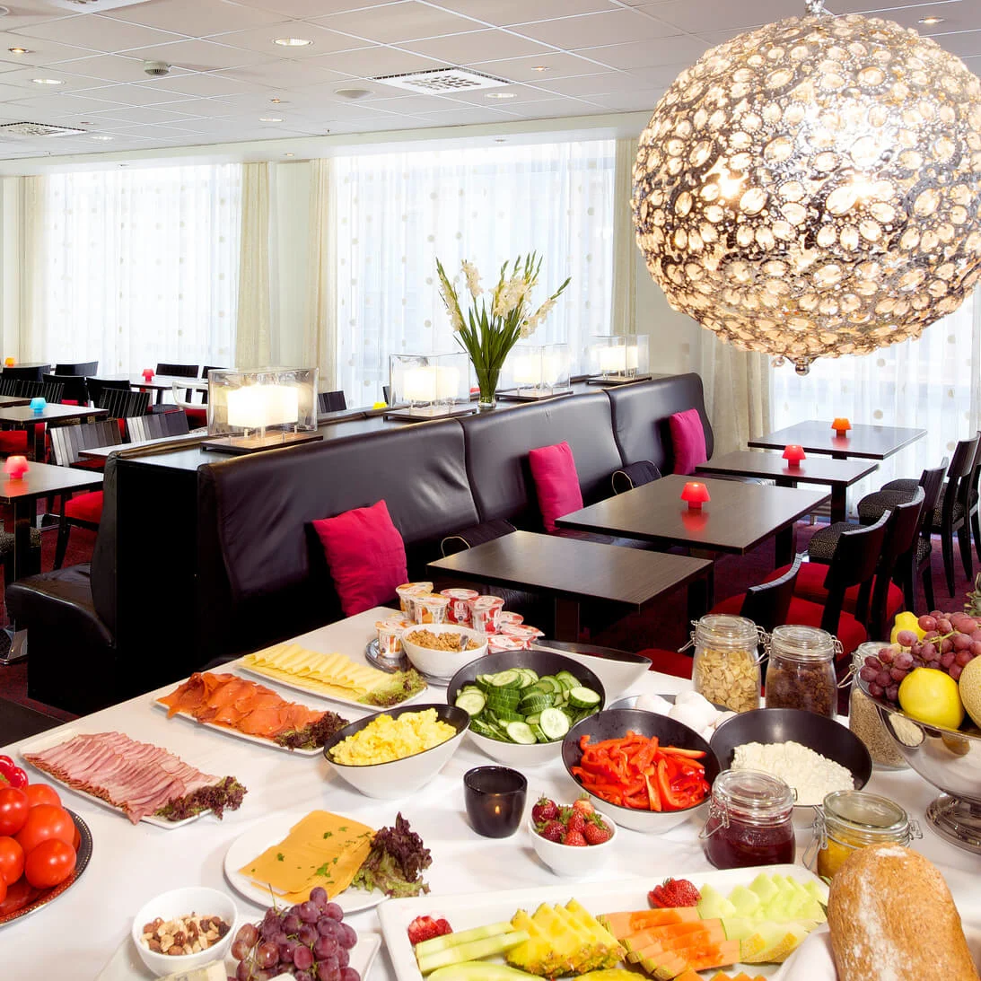 A well-lit buffet spread with various foods in a modern dining area with colorful seating and large, ornate hanging light.