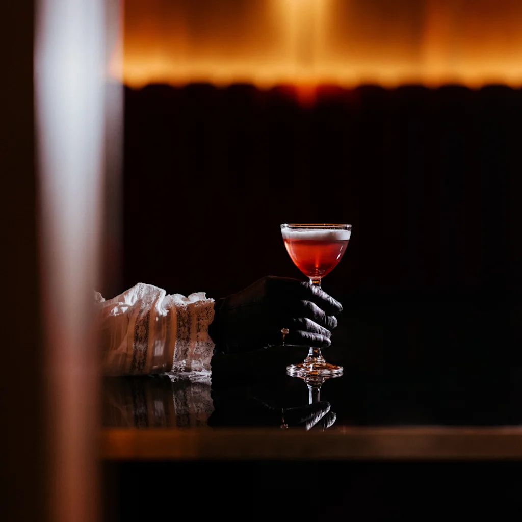 En hansket hånd holder en rød cocktail i et coupeglass, med en rysjet erme synlig, mot en mørk, varm bakgrunn.