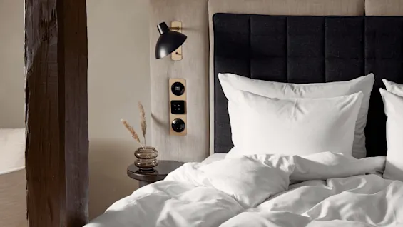 Bed with white linens and pillows, beside a wooden beam. A modern black lamp and outlet panel are mounted on the headboard. A small vase with dried grass sits on a round bedside table.