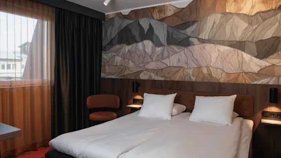 Et koselig hotellrom med en stor seng, et vindu med gardiner og en dekorativ vegg med et fjellandskapsdesign.