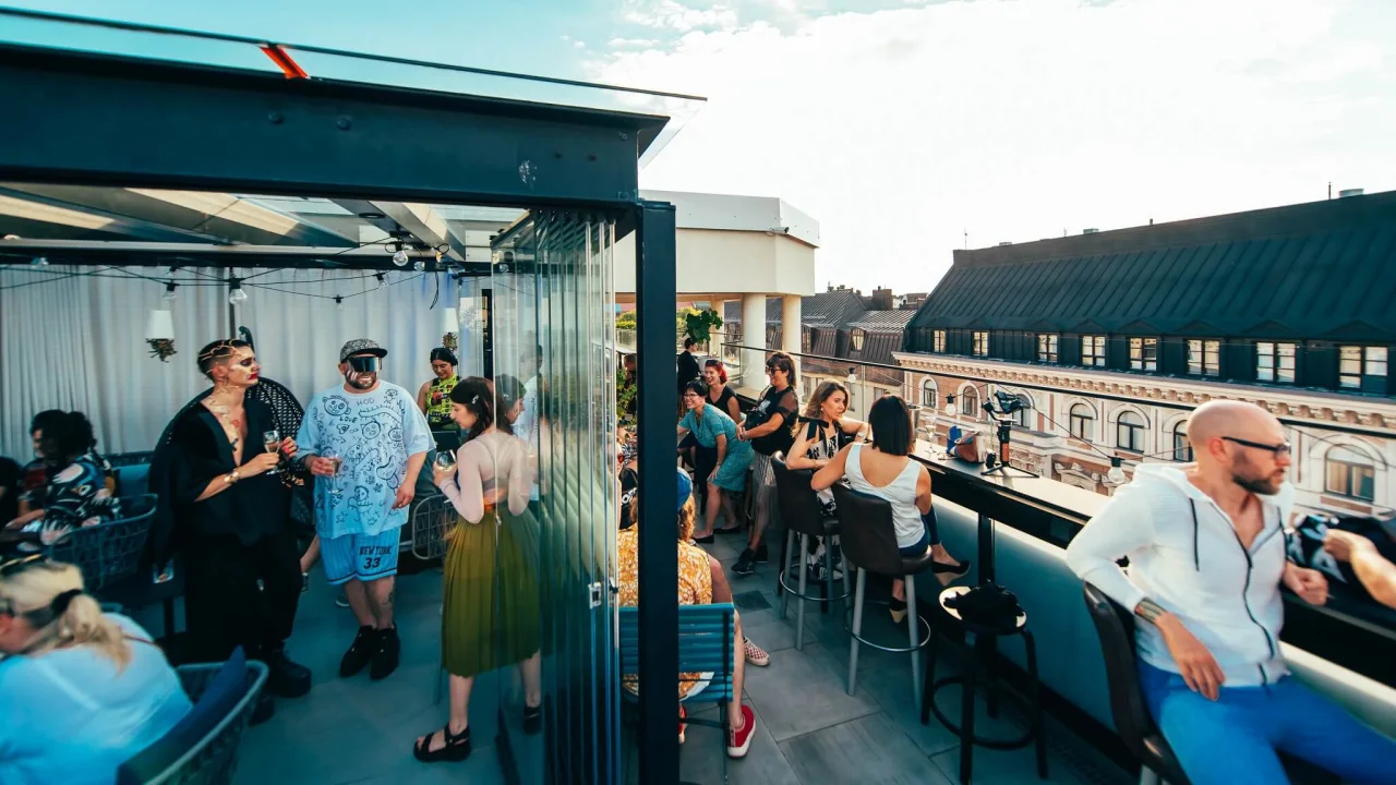 Mennesker socialiserer på en tagterrasse med drinks, bygninger i baggrunden, på en solskinsdag.