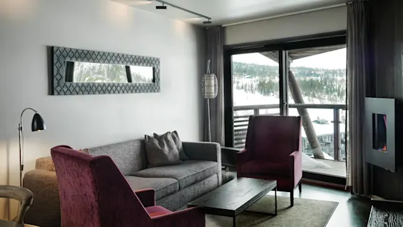A modern living room features a grey sofa, two purple armchairs, and a black coffee table; a large window reveals a snowy landscape and wooden balcony outside.