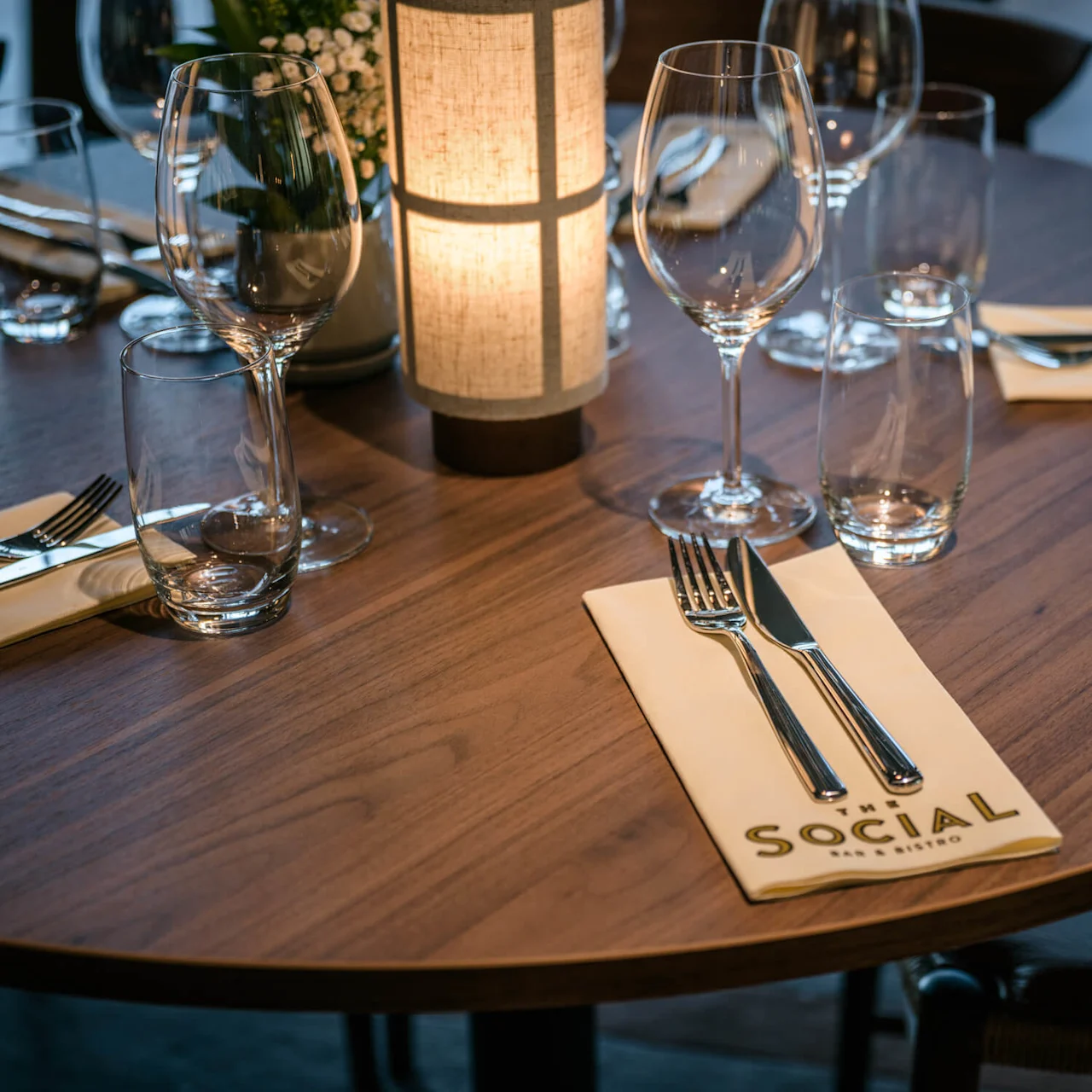 Et rundt trebord dekket for middag med vinglass, bestikk på servietter merket "THE SOCIAL BAR & BISTRO," rundt en mykt opplyst lampe. Elegant, koselig restaurantstemning.