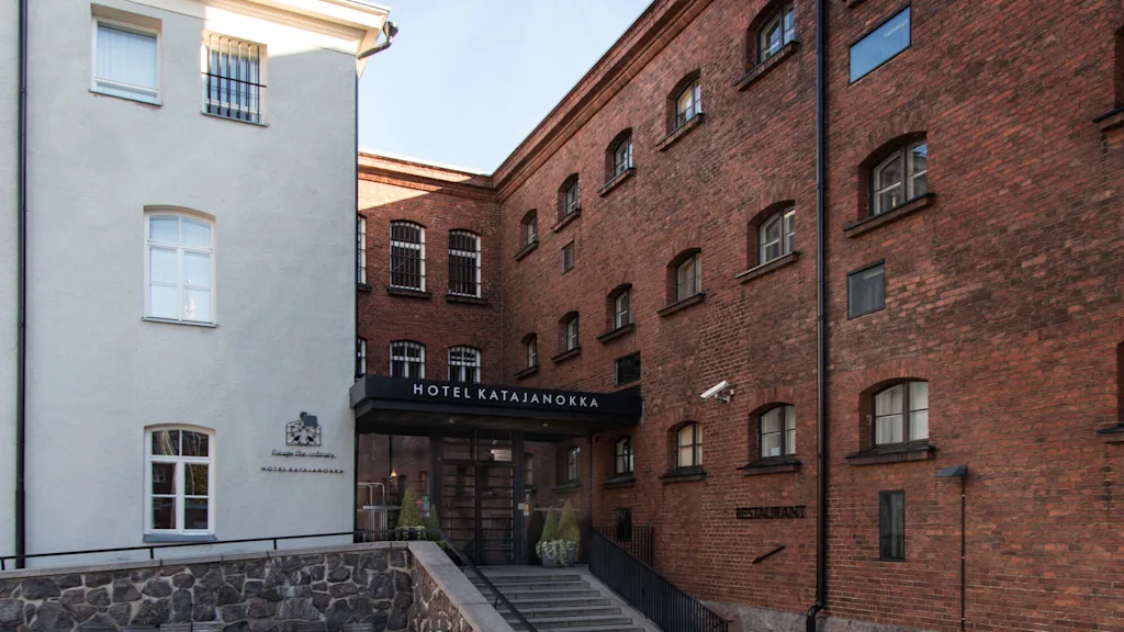 Tiilinen hotellirakennus sisäänkäyntikatoksella, kiviporrastasanteella ja viereinen valkoinen rakennus, rauhallinen sisäpiha-asetelma. Teksti: "HOTEL KATAJANOKKA" ja "Escape the ordinary".