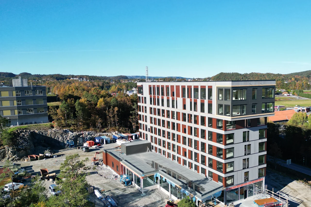 En moderne bygning under opførelse med klarblå himmel og efterårstræer i baggrunden, omgivet af byggeaktivitet.