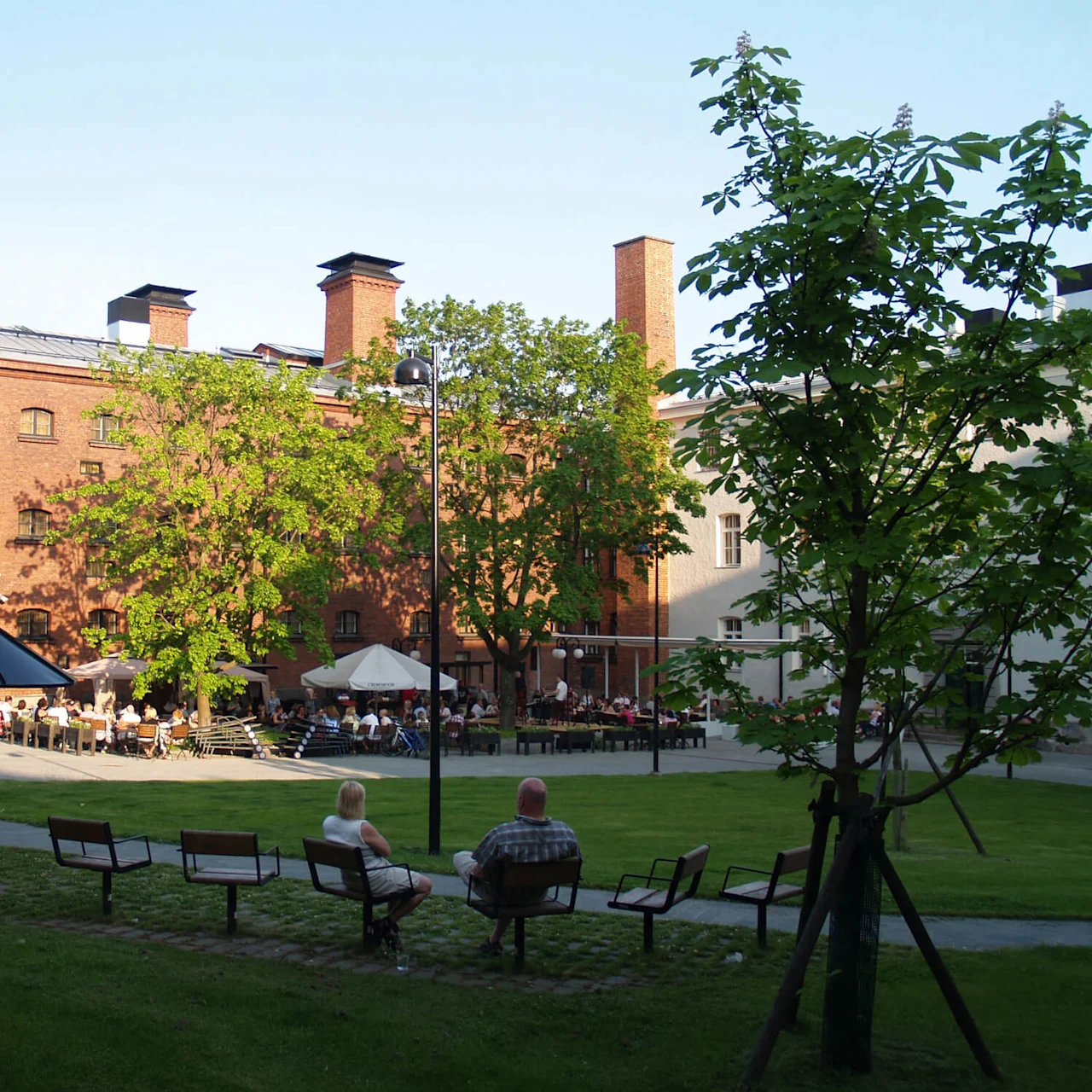 People relax at outdoor tables near a brick building with greenery, in a tranquil, urban park setting.