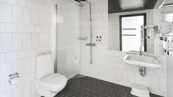 Et lyst og funktionelt hotelbadeværelse med bruser med glasvæg, toilet og håndvask med stort spejl.