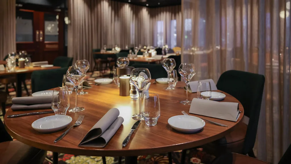 Rundt trebord dekket med vinglass, tallerkener og servietter i en elegant, svakt opplyst restaurant med polstrede stoler og gardiner.