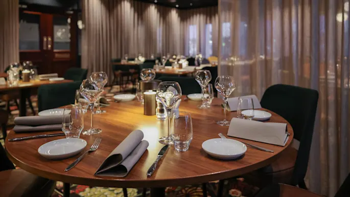 Round wooden table set with wine glasses, plates, and napkins in an elegant, dimly-lit restaurant with upholstered chairs and curtained walls.