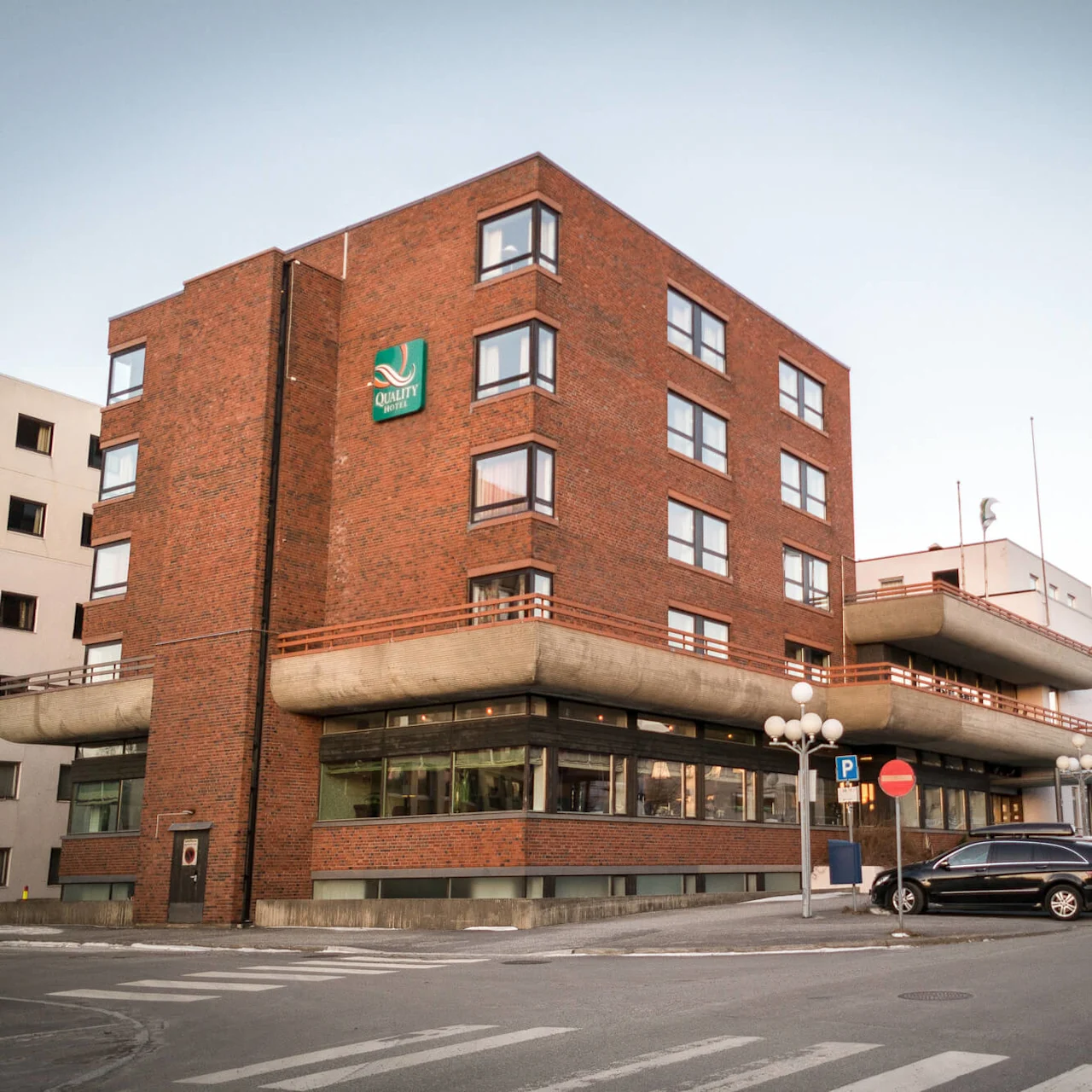 A multi-story red brick hotel building displaying a "Quality Hotels" sign, situated on a street corner, with parked cars along the sidewalk.