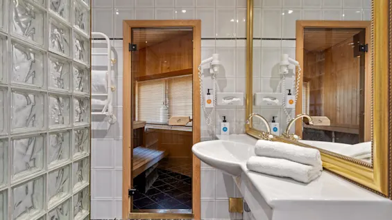 A modern bathroom features a white sink with gold faucets and folded towels, adjacent to a glass block wall. A doorway leads to a wooden sauna.