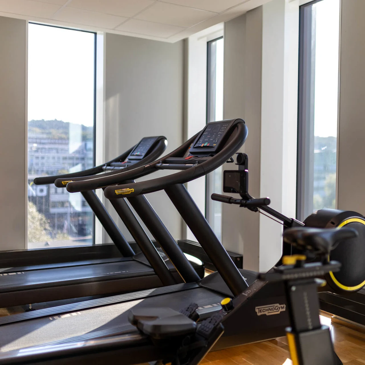 Löpband och träningscykel, placerade i ett välbelyst gym med stora fönster och trägolv. Synligt varumärke: Technogym.