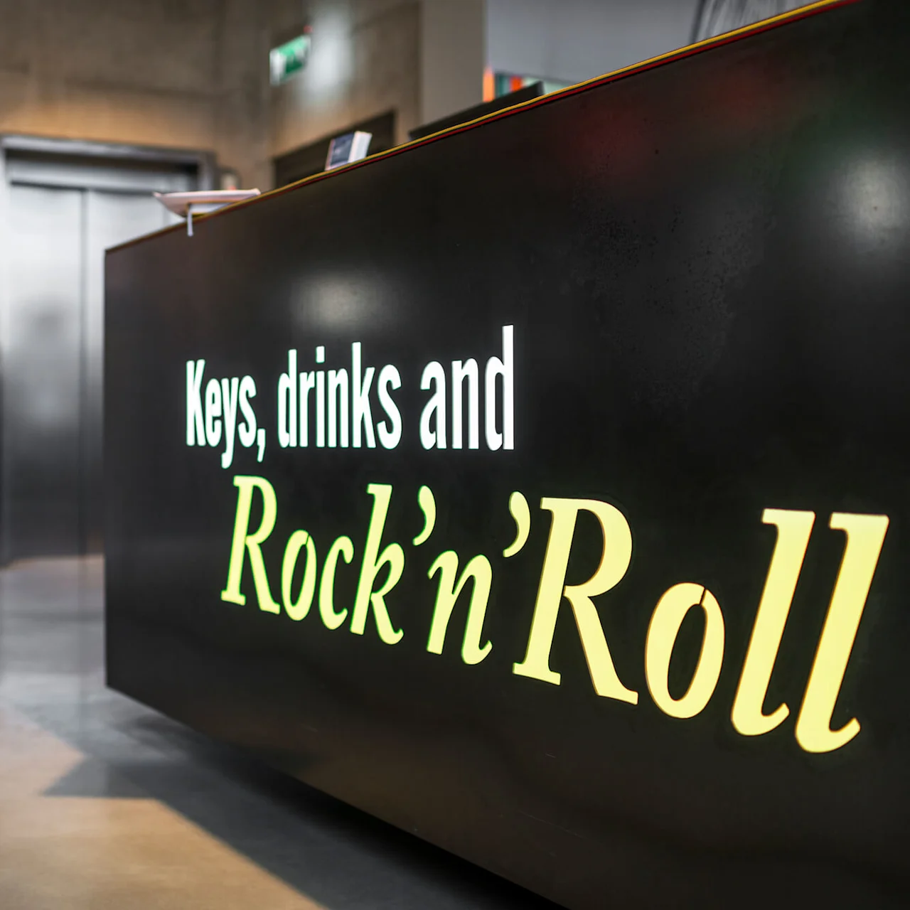 En svart disk visar texten "Keys, drinks and Rock’n’Roll" i en modern lobby, med hissar i bakgrunden.