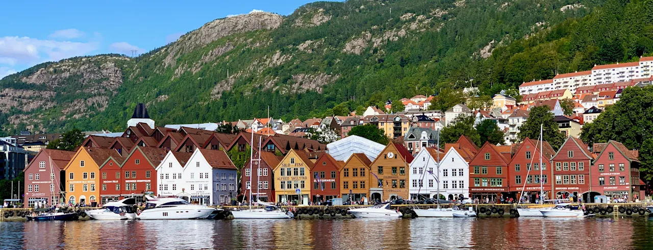 Värikkäät historialliset rakennukset Bryggenissä Bergenissä veden äärellä veneiden kanssa, vihreä vuori sinisen taivaan alla.