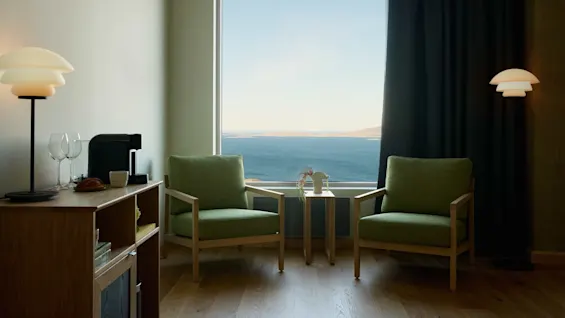 Two green armchairs face a large window overlooking the ocean. A wooden side table between them holds decorative items. Nearby, a lamp and a wooden cabinet with beverages add coziness.