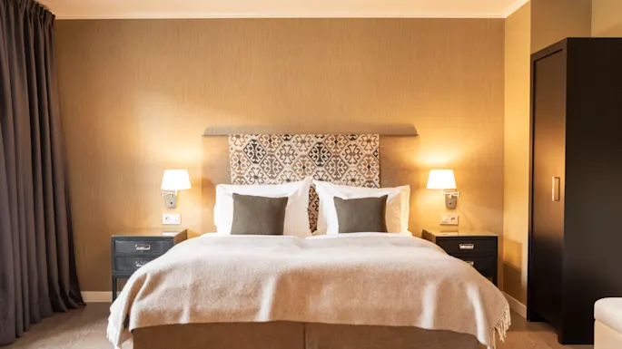 A neatly made bed with patterned headboard is flanked by two bedside tables with lamps, set in a warmly lit, contemporary-styled bedroom featuring neutral tones and a large wardrobe.