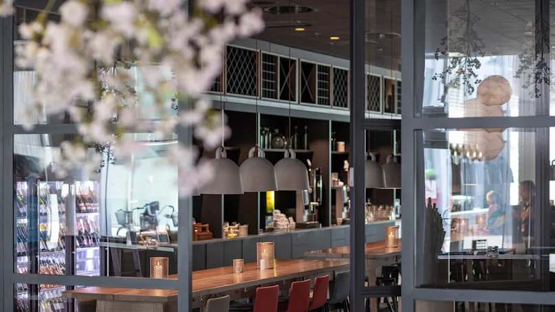 Long wooden table with red and black chairs in a modern cafe, illuminated by pendant lamps. Glass walls reveal fridges and shelves stocked with items in the background.