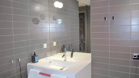 A modern bathroom with a white sink and chrome faucet. Two towels hang on a rack. Grey tiles cover the walls, reflecting soft lighting. 