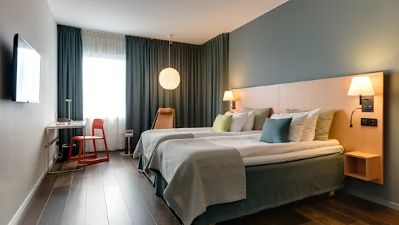 To senger er pent plassert i et moderne hotellrom, ledsaget av en nattlampe, en veggmontert TV og et skrivebord ved vinduet med gardiner.  