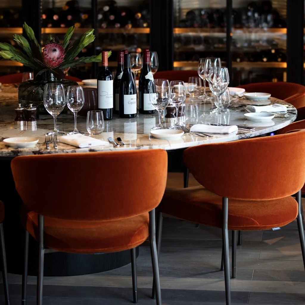 Ett elegant dukat matbord visar vinflaskor och glas, omgiven av lyxiga orange stolar i en svagt upplyst restaurang. En mittpunkt med blommor tillför dekorativ stil.