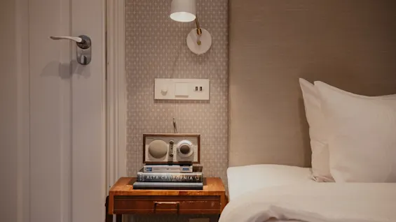 Home Hotel Wellington. Et hyggeligt hotelværelse med seng, sengebord, radio, bøger og en væglampe. ALTA CALIFORNIA.