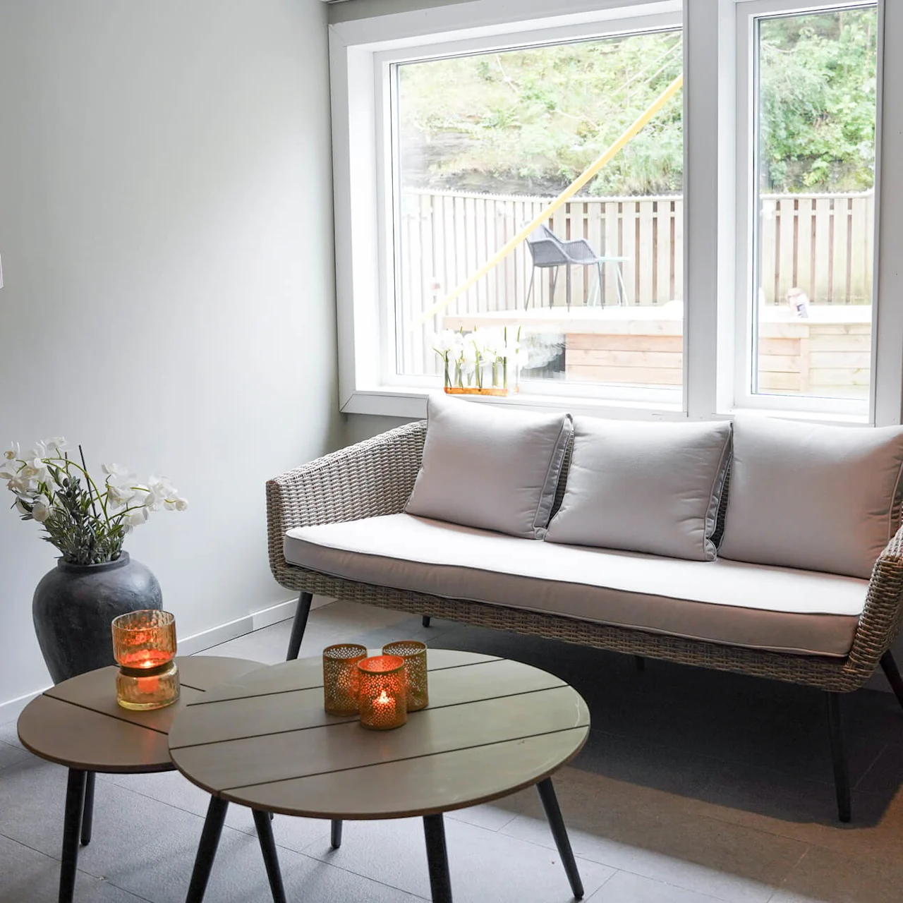 A wicker sofa with white cushions sits by a window in a cozy living room. A round table with lit candles and a vase of flowers nearby enhances the warmth.