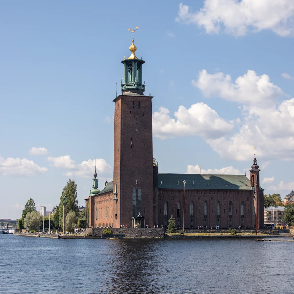 Stockholm rådhus med sit ikoniske teglstenstårn ved vandet under en blå himmel med lette skyer.