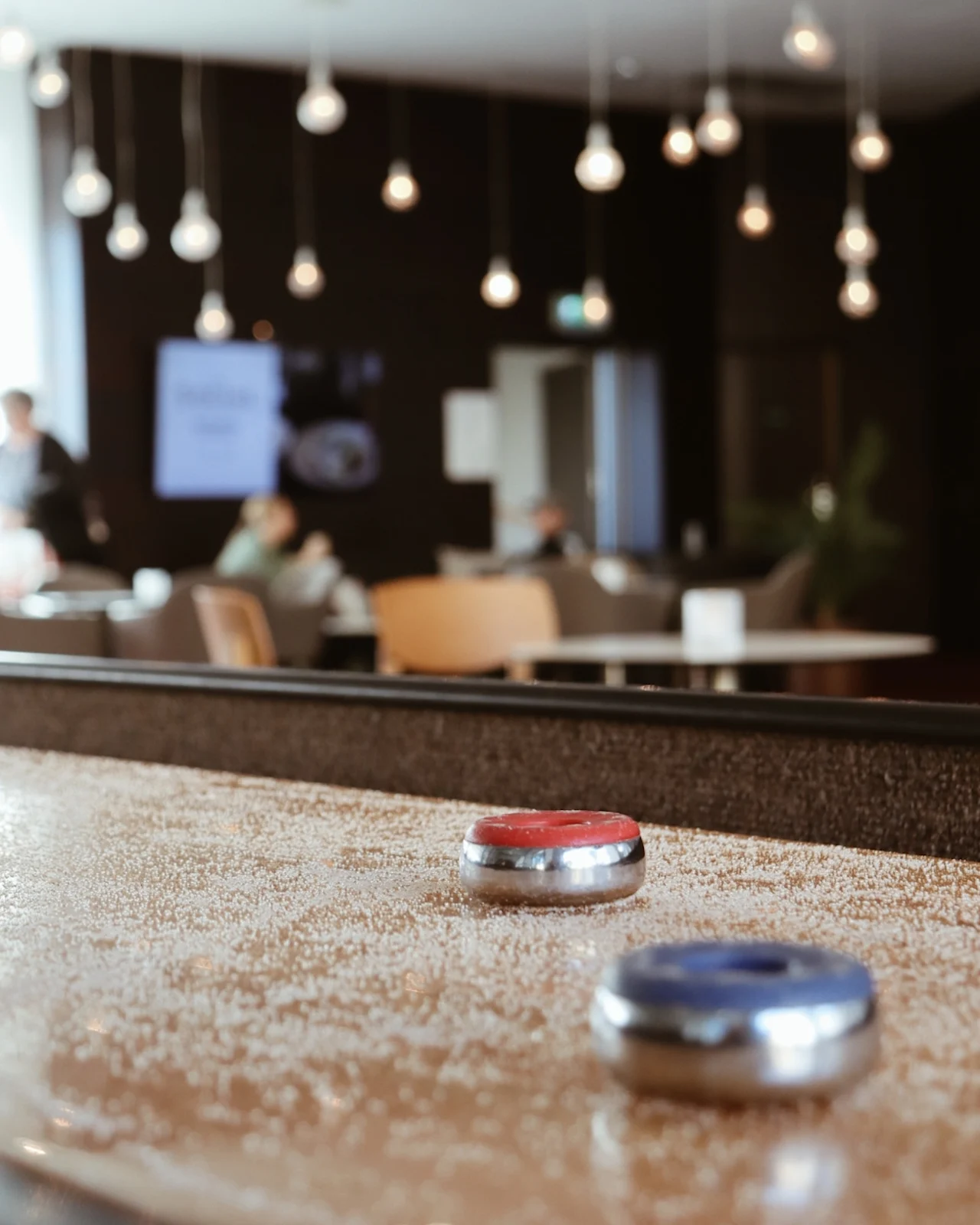 A shuffleboard with red and blue pucks on a sprinkled surface is in focus, set in a cozy lounge area with soft seating and overhead hanging lights.  