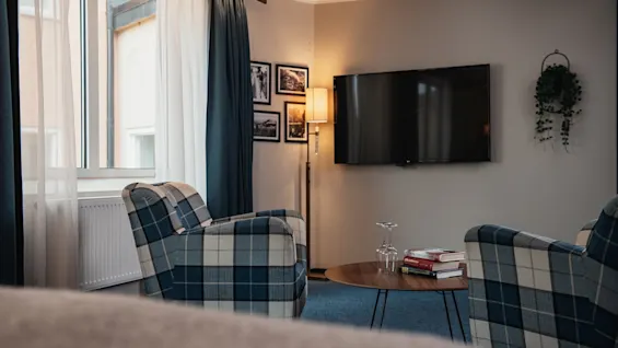 Viihtyisä hotellihuone ruudullisilla nojatuoleilla, televisiolla ja pienellä pöydällä, jossa on kirjoja, kutsuen rentoutumaan.