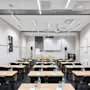 Classroom setup with wooden desks, black chairs, and bottled water. A projector screen hangs at the front, surrounded by speakers. Concrete walls display minimal posters.
