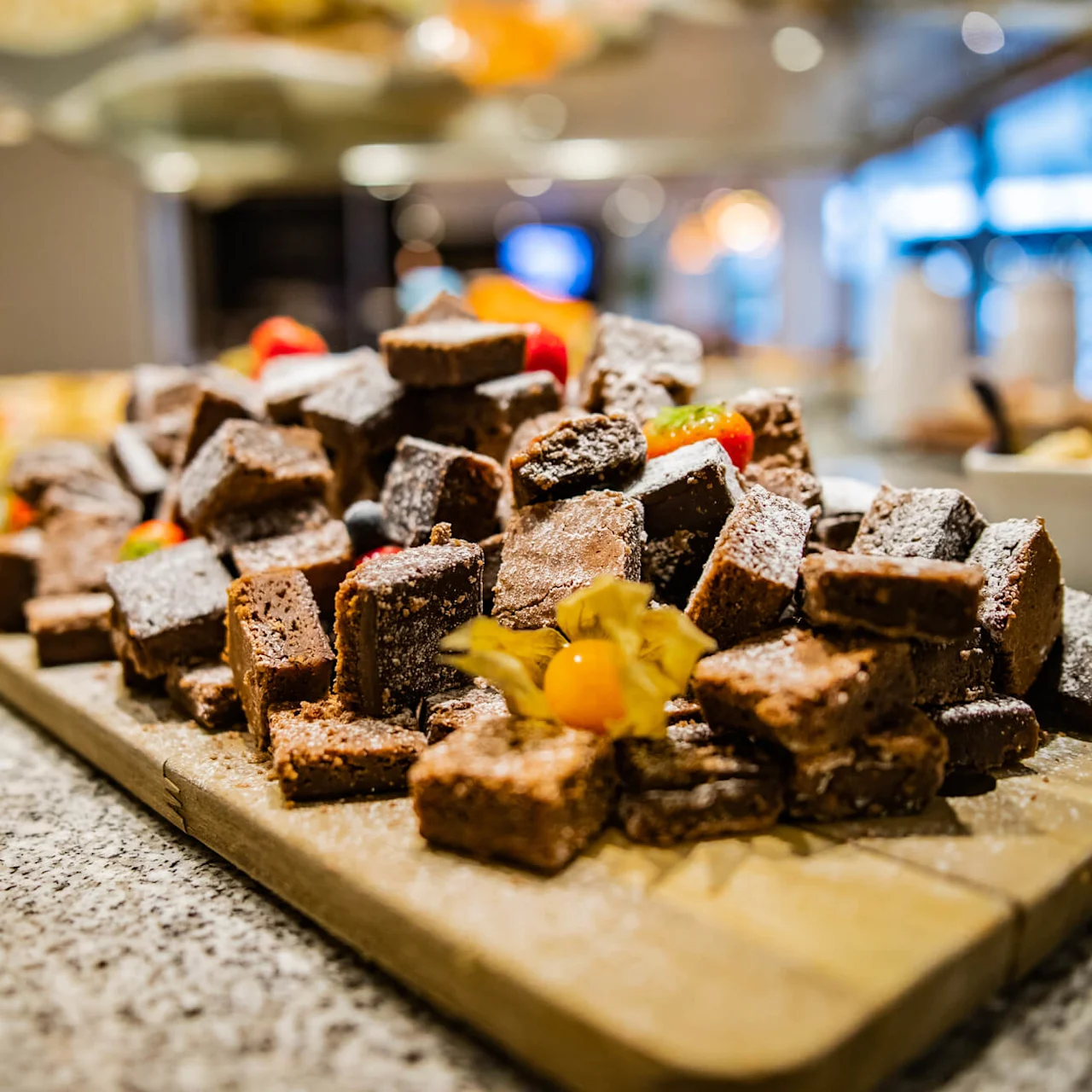 Browniebiter stablet på et brett, pyntet med melis, jordbærskiver og en physalis, i en diskret opplyst serveringsmiljø med uskarp bakgrunn.